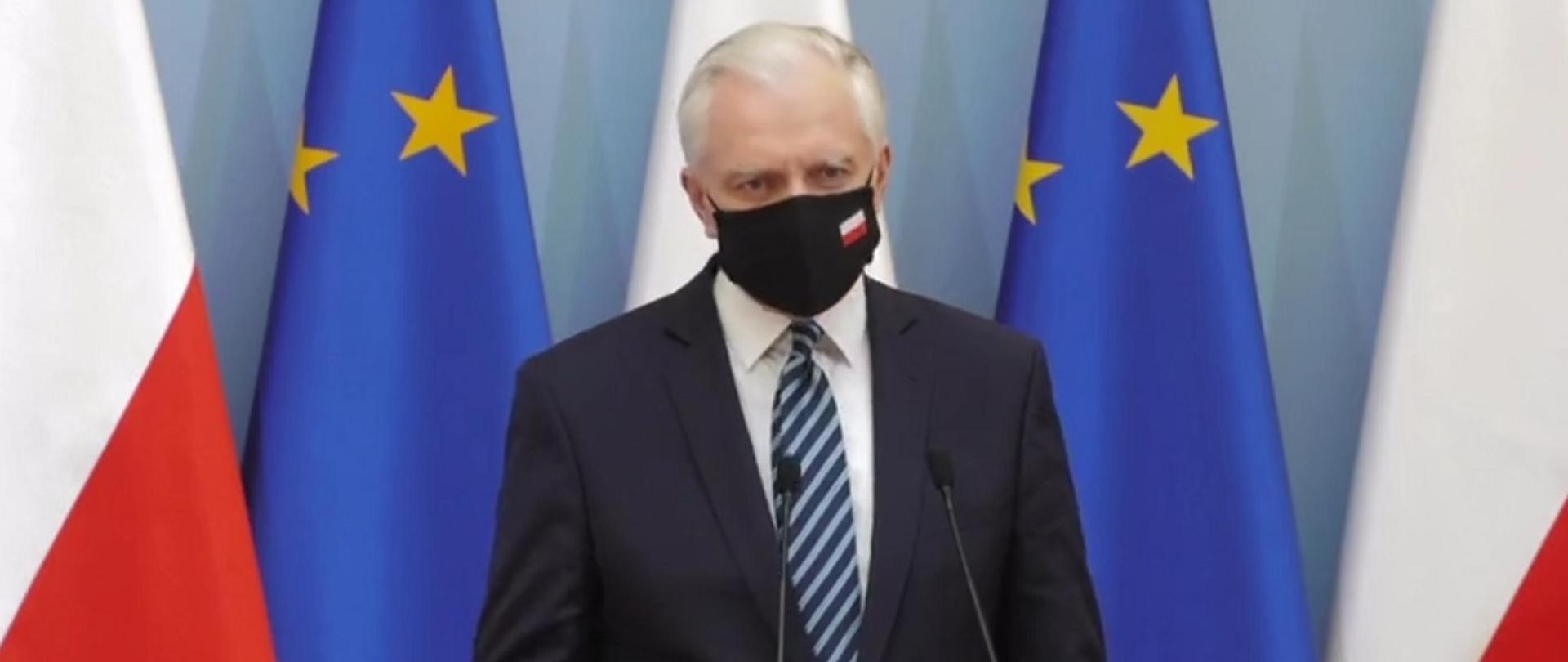 Wicepremier, minister rozwoju, pracy i technologii Jarosław Gowin stojący przy mikrofonach, za jego plecami flagi Polski i UE