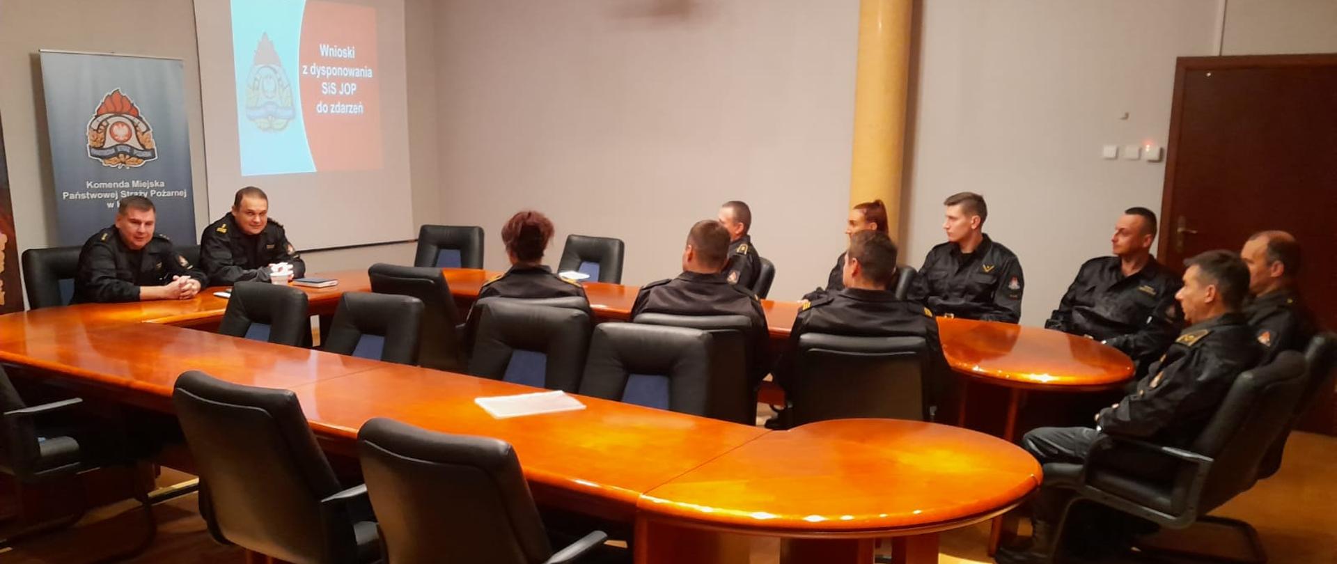 Na zdjęciu widać funkcjonariuszy KM PSP w Kaliszu. Ubrani są w umundurowanie dowódczo-sztabowe koloru czarnego. Osoby siedzą przed stołem. Zwróceni są w stronę przewodniczącego spotkania. Stoły koloru brązowego. Pomieszczenie oświetlone sztucznym światłem. Podłoga z paneli podłogowych.