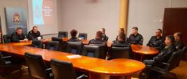 Na zdjęciu widać funkcjonariuszy KM PSP w Kaliszu. Ubrani są w umundurowanie dowódczo-sztabowe koloru czarnego. Osoby siedzą przed stołem. Zwróceni są w stronę przewodniczącego spotkania. Stoły koloru brązowego. Pomieszczenie oświetlone sztucznym światłem. Podłoga z paneli podłogowych.