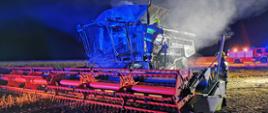 Zdjęcie przedstawia doszczętnie spalony kombajn zbożowy marki Class, znajdujący się na spalonym ściernisku. W tle znajdują się samochód ratowniczo-gaśniczy oświetlający teren akcji. Zdjęcie wykonano nocą.