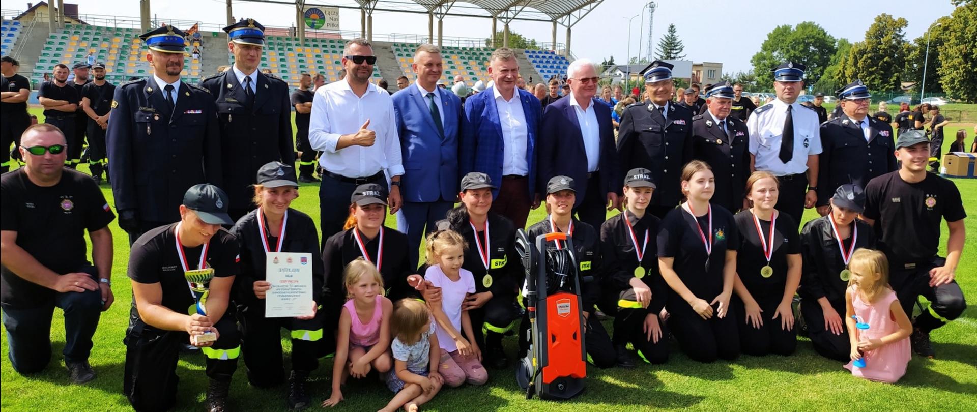 Kobieca drużyna OSP pozuje do zdjęcia na stadionie piłkarskim, stoji z nimi również kilku druhów i strażaków w mundurach wyjściowych oraz kilku mężczyzn w marynarkach