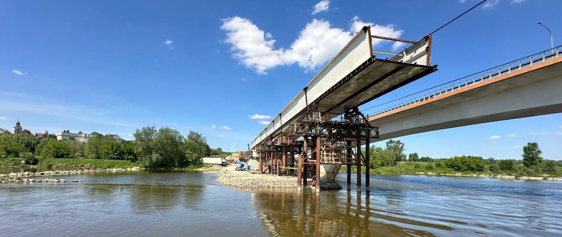 Budowa mostu w Sandomierzu - nad rzeką most użytkowany po prawej i budowany po lewej, podłużny element konstrukcji z przymocowanymi z przodu linami oparty na podporach tymczasowych, w oddali za drzewami budynki starego miasta 