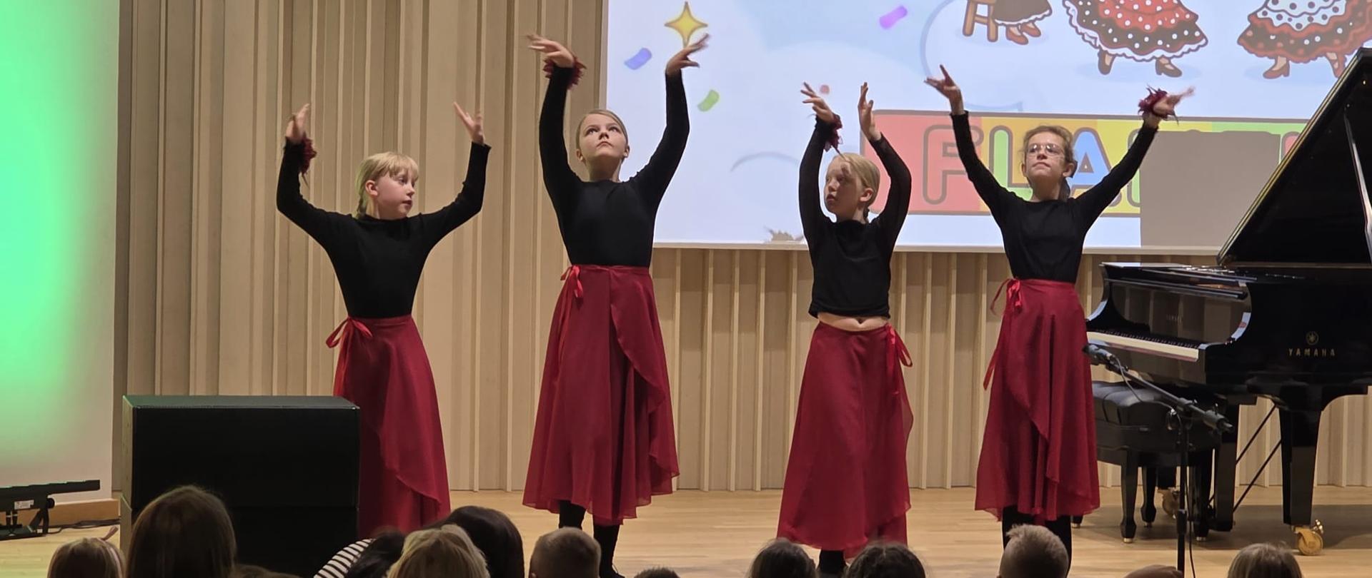 Na zdjęciu cztery dziewczyny w czarnych bluzkach i czerwonych spódnicach, które tańczą na brązowej scenie. Obok nich czarny fortepian. Przed sceną licznie zgromadzona publiczność.