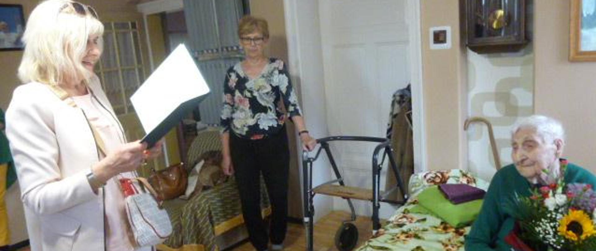 Stojąca w pokoju kobieta odczytuje siedzącej na łóżku i trzymającej bukiet kwiatów Jubilatce list gratulacyjny. W głębi pokoju stoi inna kobieta.