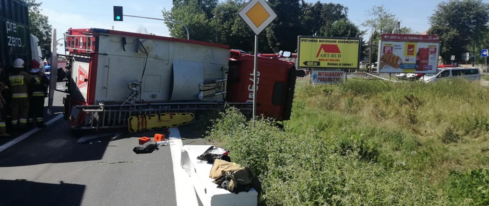 
Zdjęcie przedstawia przewrócony na prawym boku samochód pożarniczy po zderzeniu z samochodem osobowym, pojazd częściowo blokuje pas jezdni.