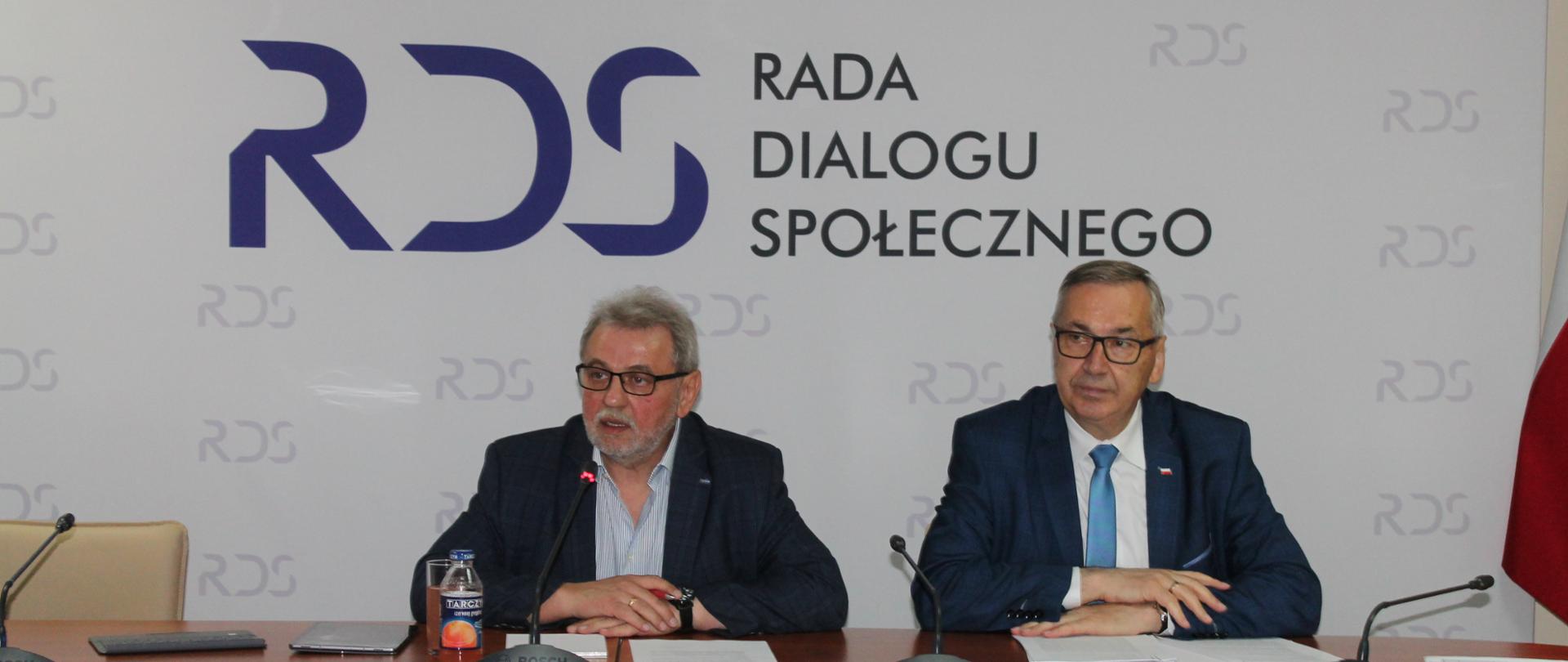 Na zdjęciu znajduje się stół prezydialny za którym siedzą od lewej strony: Henryk Nakonieczny, Przewodniczący Zespołu problemowego ds. prawa pracy a następnie Stanisław Szwed, Sekretarz Stanu w Ministerstwie Rodziny i Polityki Społecznej. W tle siedzących osób znajduje się baner z napisem Rada Dialogu Społecznego. Każdej z osób przypisany jest mikrofon.