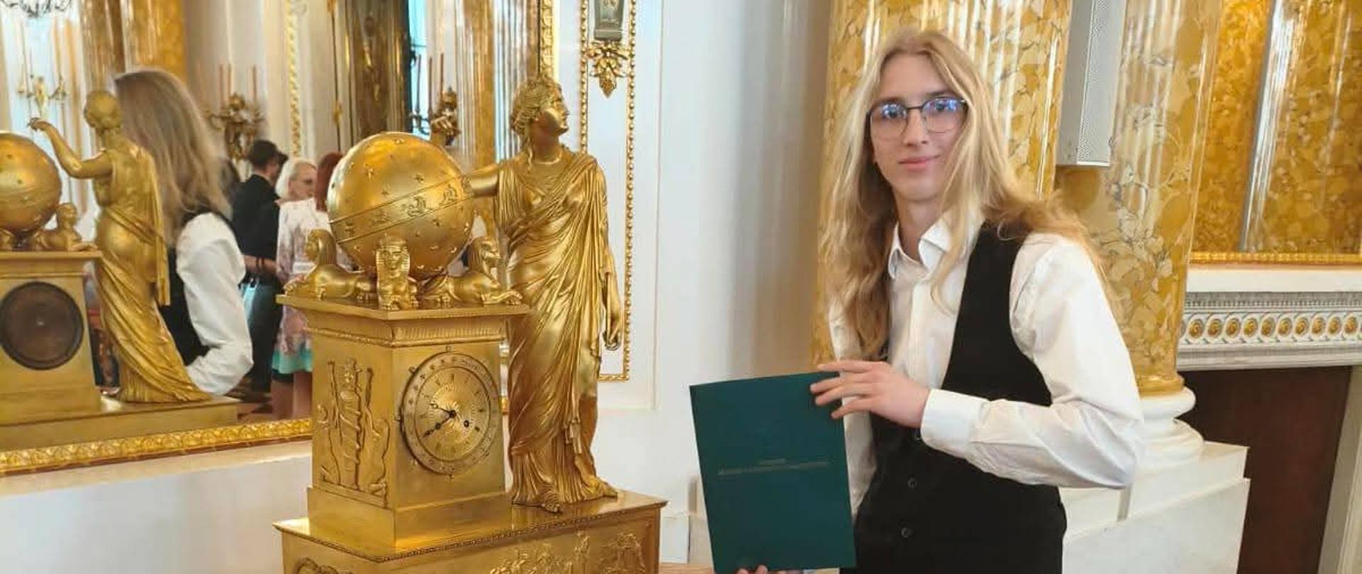 Zdjęcie w sali w Zamku Królewskiego w Warszawie sala balowa Bursztynowa złoty stół inkrustowany złoty zegar ozdobny lustro kryształowe podłoga w dwóch kolorach drewniana brązowa i w kolorze drewna przy stole stoi młody człowiek jasne długie blond włosy rozpuszczone okulary czarna kamizelka biała koszula czarne spodnie trzyma w dłoniach zieloną teczkę zapewne z dyplomem potwierdzającym otrzymanie stypendium Ministra Kultury i Dziedzictwa Narodowego