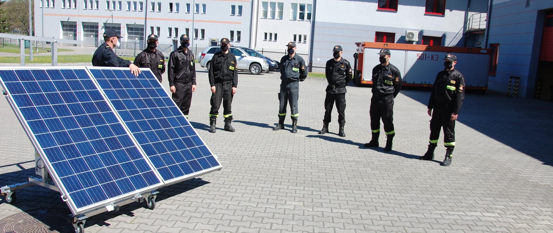 Na pierwszym planie zdjęcia widać dwa panele fotowoltaiczne skierowane przodem w kierunku słońca. Za panelami stoi instruktor, obok 7 strażaków ubranych w ubrania koszarowe. Strażacy na głowach czapki z daszkiem. Wszyscy ludzie na zdjęciu mają na twarzach maseczki. Plac na którym odbywają się zajęcia jest wybrukowany kostką. W tle zdjęcia widać budynek strażnicy.