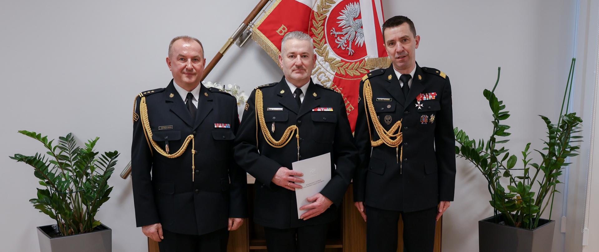 Trzech strażaków w mundurach wyjściowych ze sznurem stoi obok siebie strażak w środku trzyma białą teczkę za nim na ścianie umieszczony na uchwytach jest sztandar Komendy Głównej Państwowej Straży pod nim jest komoda z figurką oraz po obu stornach szafki ustawione są kwiaty w donicach.