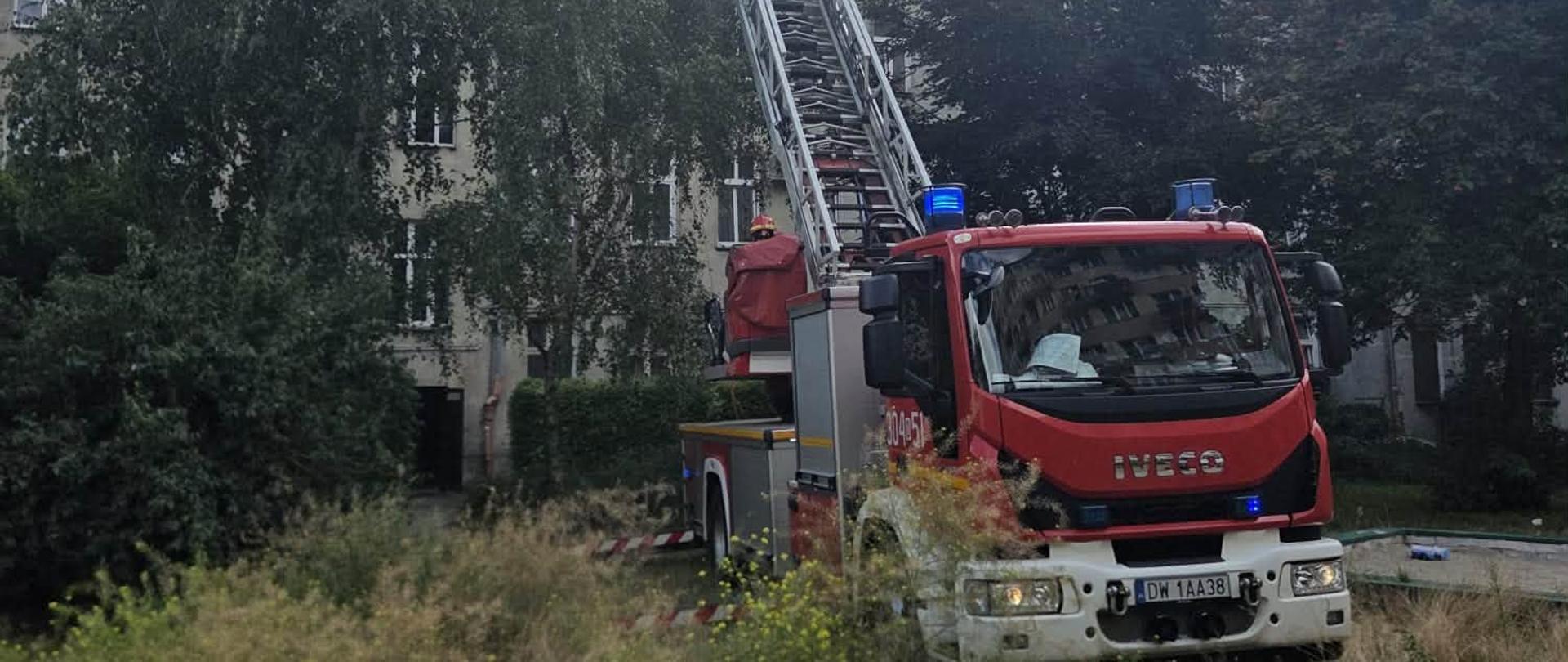 Samochód Specjalny Drabina (SD) rozstawiony na tle budynku mieszkalnego wielorodzinnego z wysuniętym koszem drabiny do ostatniego piętra. 