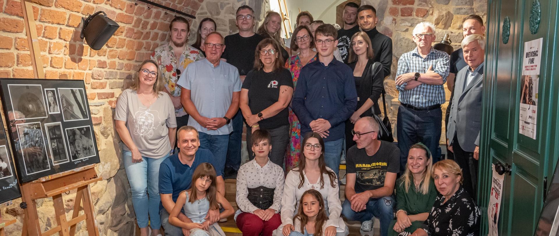Zdjęcie przedstawia publiczność zebraną podczas wernisażu wystawy fotograficznej. Grupa osób pozuje na tle ceglanych ścian.