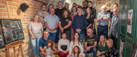 Zdjęcie przedstawia publiczność zebraną podczas wernisażu wystawy fotograficznej. Grupa osób pozuje na tle ceglanych ścian.