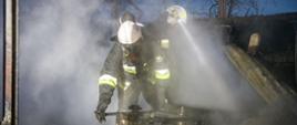 Strażacy OSP w ubraniach specjalnych, aparatach ochrony układu oddechowego oraz białych hełmach działają podczas dogaszania pożaru wiaty magazynowej na tartaku w Prószkowie. Strażacy pracują w widocznym zadymieniu. 