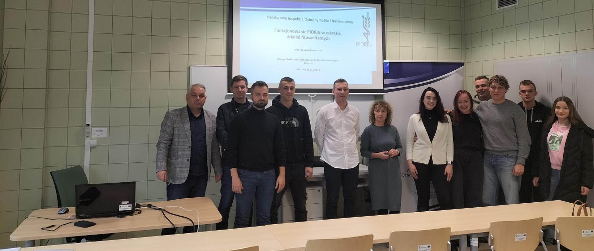 Zdjęcie przedstawia jasną, dużą salę wykładową ze stołami i krzesłami. W jej centralnym miejscu znajduje się duży ekran, na którym widnieje tekst: Funkcjonowanie PIORiN w zakresie działań fitosanitarnych. Przy ścianie stoi grupa dziewięciu studentów (trzy kobiety i sześciu mężczyzn) oraz pani dziekan, wykładowca i inspektor WIORiN Kielce.