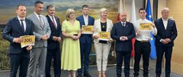 Grupa osób pozuje do zdjęcia, część trzyma tabliczki z napisem „1000 zł”, w tle flagi i baner.