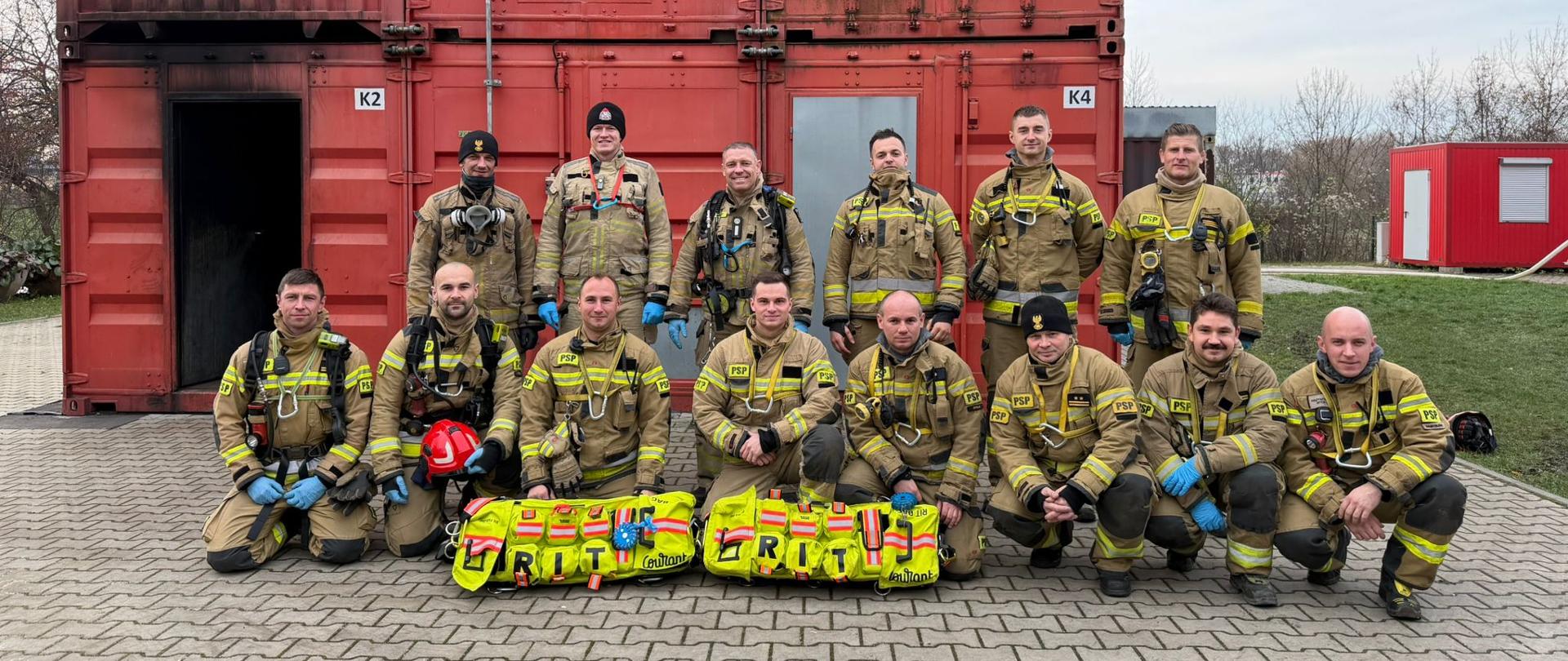 Strażacy z województwa opolskiego pozują do zdjęcia grupowego przed kontenerem treningowym oznaczonym „K2” i „K4”. Dwóch uczestników trzyma tablice z napisem „GRIT”, symbolizujące wytrwałość i zaangażowanie podczas szkolenia rot asekuracyjnych.