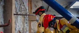 Dwóch strażaków pracuje przy uszkodzonej ścianie, używając narzędzi ręcznych. Widoczne oznaczenia sprayem.
