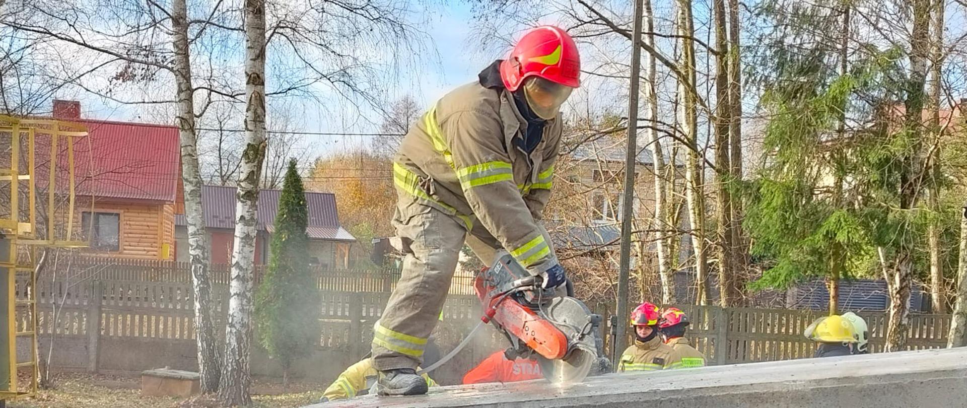 Strażak przecina piłą mechaniczną betonowy kloc, z tyłu widać innych strażaków oraz domy w tle