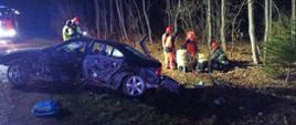 Zdjęcie przedstawia stojący na drodze i częściowo na poboczu uszkodzony samochód osobowy marki BMW. Pojazd widoczny bokiem od strony kierowcy posiada liczne uszkodzenia nadwozia. Za pojazdem po prawej strony stoją strażacy i ratownicy medyczni. Zdjęcie wykonane nocą