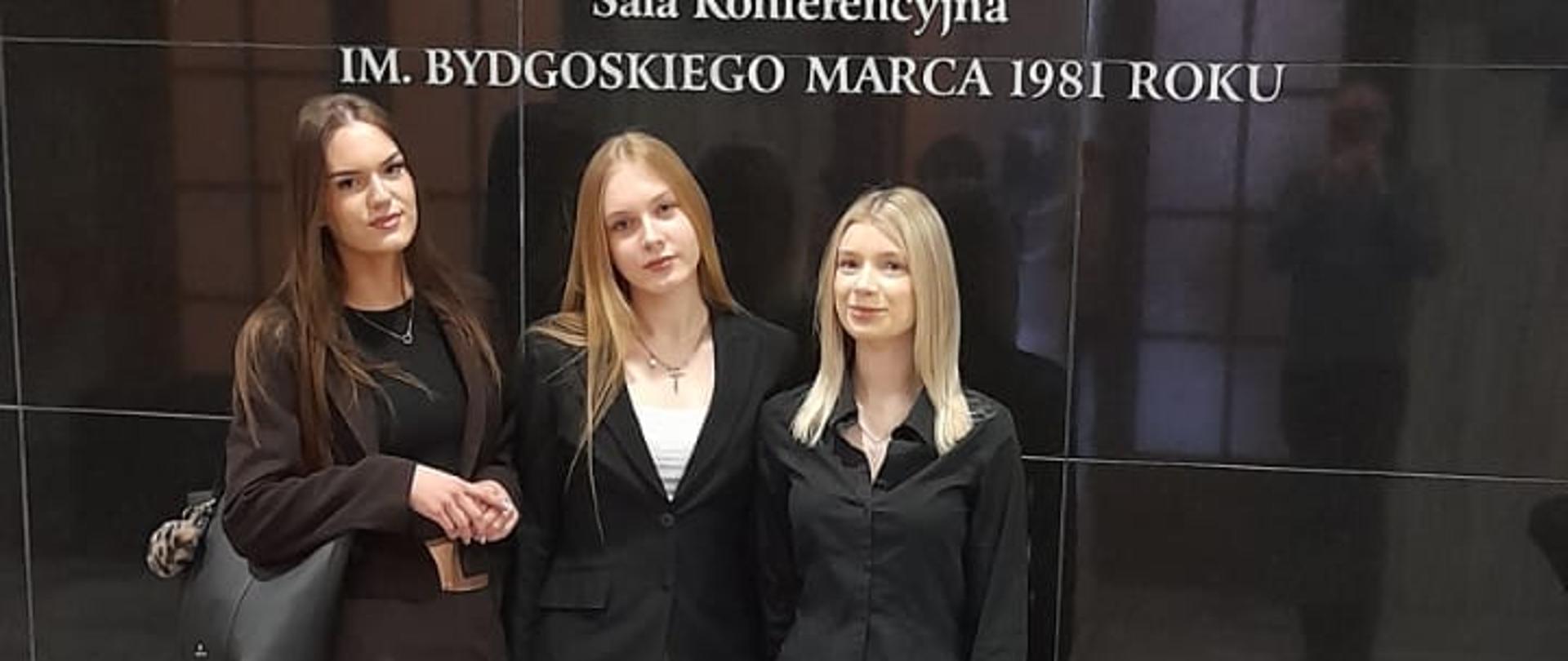 Trzy uczennice ZSCKR w Samostrzelu stoją na marmurowej podłodze w holu Kujawsko-Pomorskiego Urzędu Wojewódzkiego w Bydgoszczy. Za nimi czarna ściana z białym napisem: "KUJAWSKO-POMORSKI URZĄD WOJEWÓDZKI W BYDGOSZCZY Sala Konferencyjna IM. BYDGOSKIEGO MARCA 1981 ROKU".