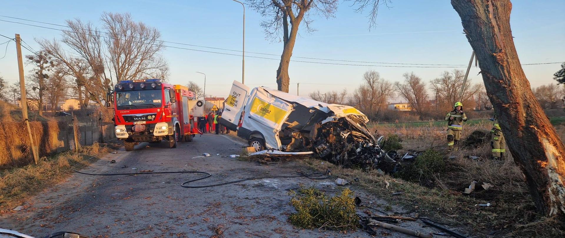 Zdjęcie przedstawia:
od lewej strony biały samochód typu bus po uderzeniu w drzewo, następnie czerwony wóz straży pożarnej