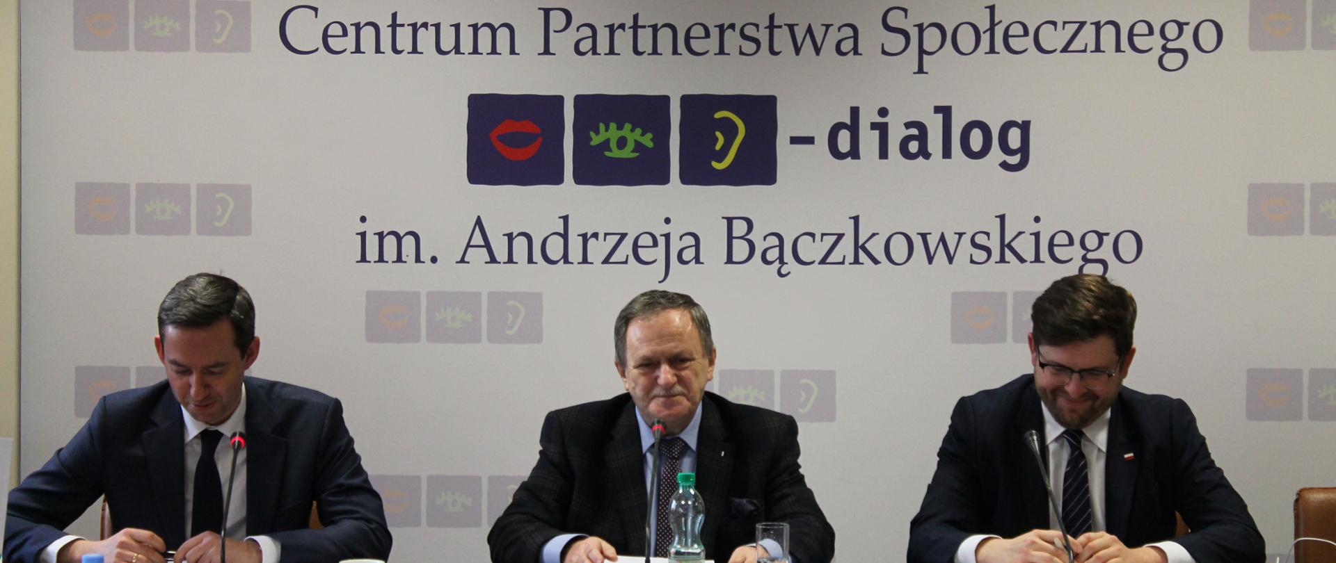 Zdjęcie przedstawia stół prezydialny, za którym siedzą: Marcin Ociepa, Sekretarz Stanu w Ministerstwie Obrony Narodowej; Andrzej Śliwka, Podsekretarz Stanu w Ministerstwie Aktywów Państwowych, Współprzewodniczący Zespołu ze strony rządowej oraz Stanisław Głowacki, Współprzewodniczący Zespołu ze strony związków zawodowych. W tle znajduje się baner z napisem Centrum Partnerstwa Społecznego „Dialog” im. Andrzeja Bączkowskiego.