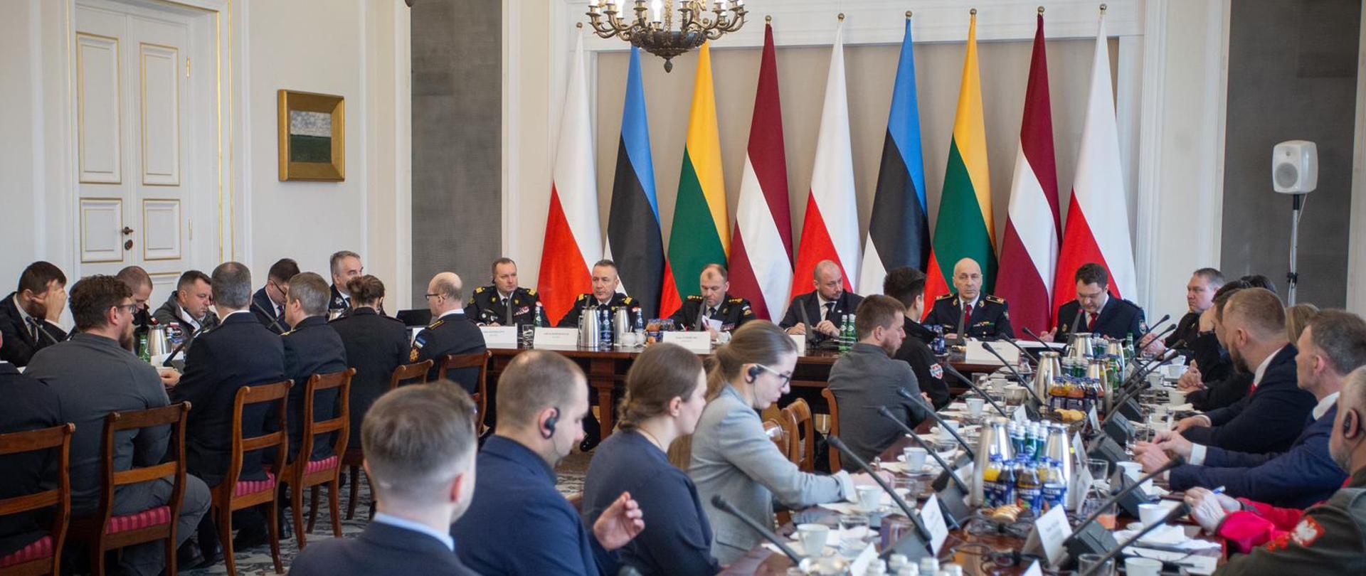 Przy stołach podczas konferencji siedzą jej uczestnicy, za frontowym stołem stoją flagi Polski, Estonii, Litwy, Łotwy