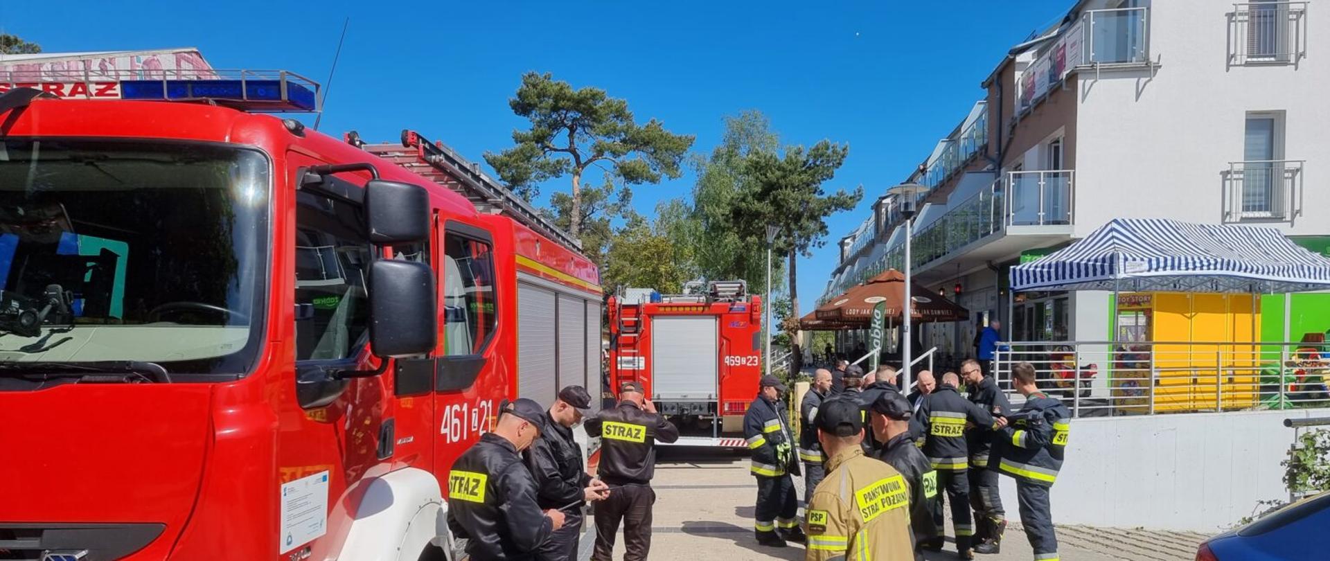 Zdjęcie przedstawia wozy strażackie oraz strażaków na ulicy .W tle widać budynek oraz drzewa.