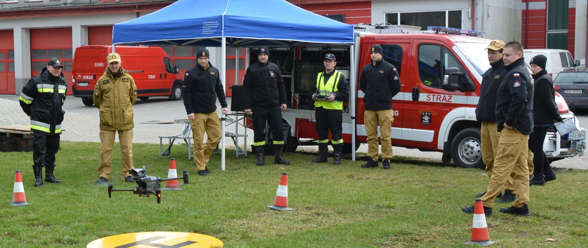 strażacy przyglądają się startującemu dronowi za nimi mały namiot konferencyjny z niebieskim dachem oraz samochód strażacki w tle budynek komendy straży