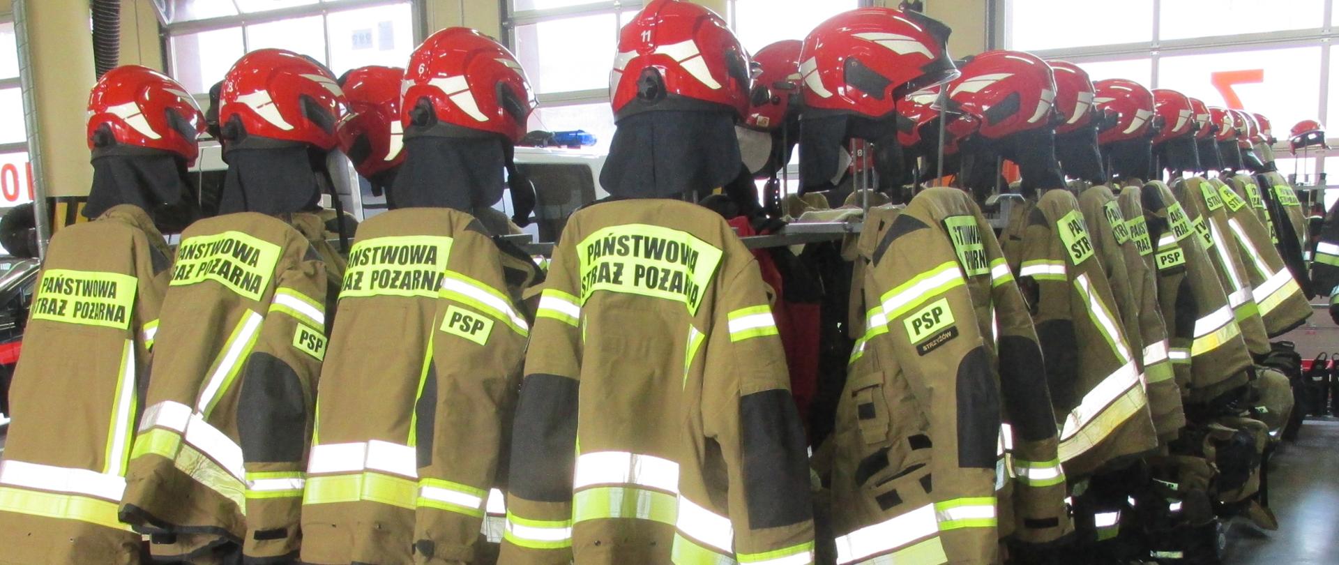 Na zdjęciu widoczne ubrania specjalne w kolorze żółtym. Kurtki wiszą na wieszaku znajdującym się na garażu jednostki ratowniczo-gaśniczej. Pod kurtkami znajdują się spodnie naciągnięte na obuwie specjalne. Nad kurtkami widoczne nowe hełmy strażackie w kolorze czerwonym. Zdjęcie wykonano w sposób ukazujący dwa rzędy ubrań.