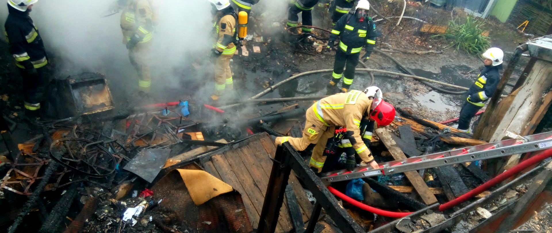 Na zdjęciu widać strażaka, który wchodzi po drabinie na poddasze uszkodzonego budynku - w tle znajdują się strażacy przeszukujący zgliszcza.