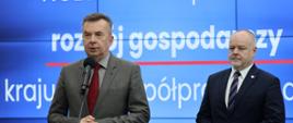 Na tle wbudowanego w ścianę wielkiego ekranu z napisem NCBR pod nadzorem Ministra Nauki stoi minister Wieczorek, który mówi do mikrofonu na stojaku, obok niego wiceminister Gzik.