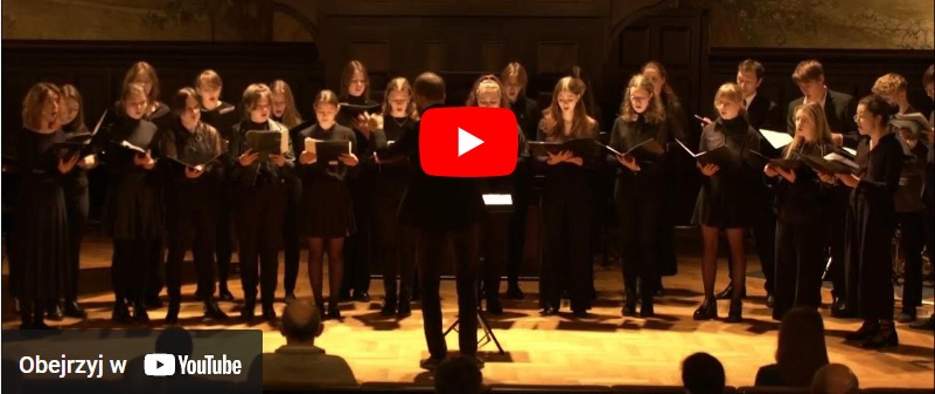Koncert PSM w Akademii Muzycznej w Katowicach - zdjęcie przedstawia zrzut z odtwarzacza YouTube, na grafice znajduje się chór mieszany, który śpiewa na scenie