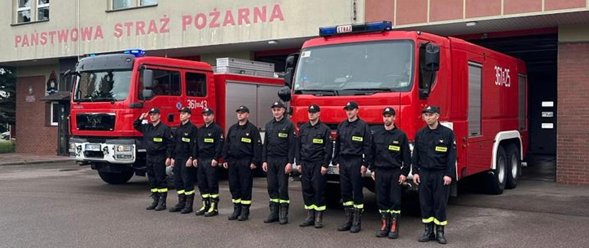 Strażacy oddający honor w tle widoczne 2 samochody pożarnicze i budynek komendy.