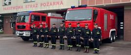 Strażacy oddający honor w tle widoczne 2 samochody pożarnicze i budynek komendy.