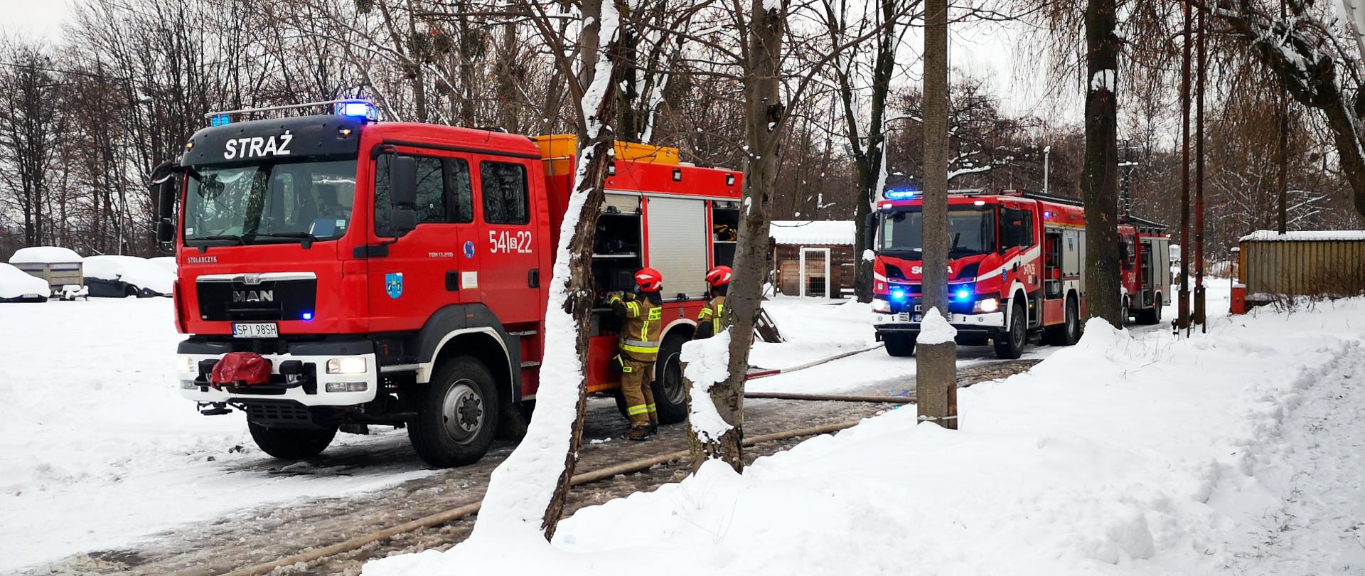 Zdjęcie przedstawia trzy czerwone samochody ratowniczo - gaśnicze z włączonymi światłami ostrzegawczymi stojące na zaśnieżonej drodze. Obok pierwszego samochodu widać dwóch strażaków wyciągających ze skrytek sprzęt ratowniczy. Z tyłu za samochodami widać drzewa.