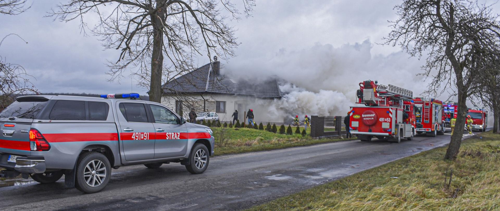 Zdjęcie przedstawia pożar budynku jednorodzinnego, wydobywający się dym ogranicza widoczność i utrudnia działania gaśnicze. Strażacy podawają dwa prądy wody w natarciu na palący się obiekt.