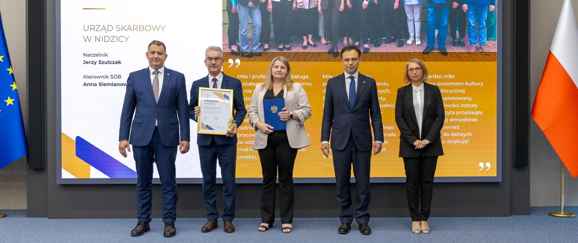 Andrzej Domański, minister finansów i gospodarki, Marcin Łoboda, szef Krajowej Administracji Skarbowej, oraz przedstawiciele Urzędu Skarbowego w Nidzicy prezentują dyplom i statuetkę „Gwiazda Jakości". Na ekranie w tle widoczna jest nazwa urzędu, zdjęcie zespołu oraz fragmenty opinii klientów.