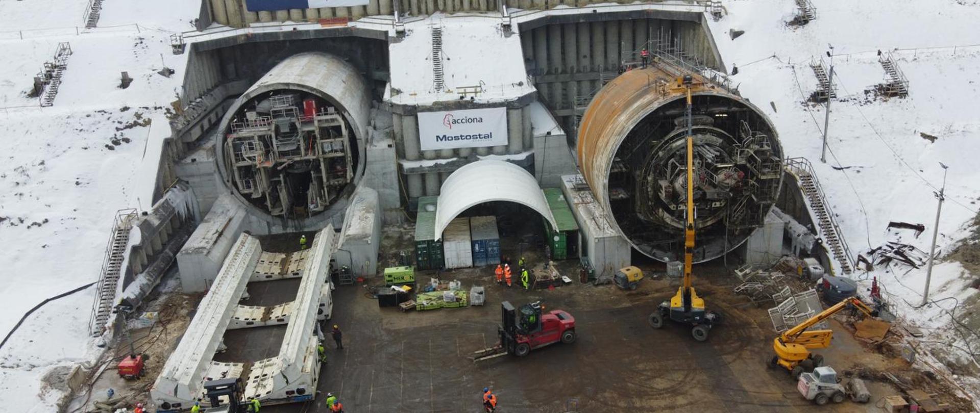 Na zdjęciu widać duży, zimowy plac budowy tunelu drogowego. Przed ścianą wykutą w zboczu znajdują się dwa ogromne, okrągłe otwory tunelowe. W każdym otworze stoi maszyna TBM – po lewej tarcza jest odsłonięta, a po prawej widać duży, brązowy, cylindryczny korpus. Przed nimi pracuje kilka żółtych i czerwonych maszyn budowlanych, a na ziemi poruszają się robotnicy w jaskrawych, odblaskowych ubraniach. Wokół placu znajduje się śnieg, a całość sprawia wrażenie intensywnych prac przy dużej inwestycji tunelowej.