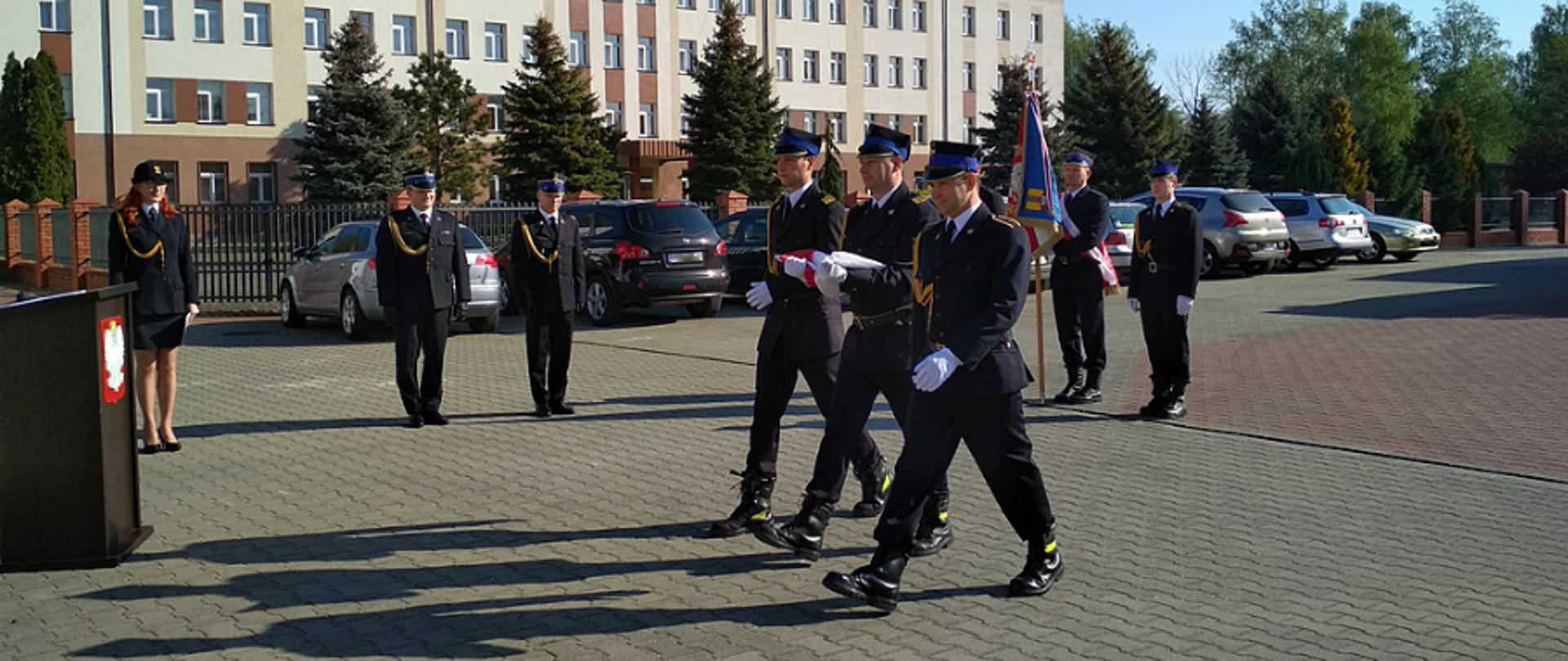 na zdjęciu trzyosobowy poczet flagowy maszeruje na placu wewnętrznym komendy powiatowej w Łowiczu w stronę masztu flagowego, strażacy ubrani są w mundury galowe ze sznurem, na głowie maja czapkę rogatywkę i białe rękawiczki, strażak idący w środku trzyma w dłoniach złożoną flagę państwową, po lewej stronie znajduje się mównica, w tle stoją funkcjonariusze komendy, poczet sztandarowy oraz komendant powiatowy mł. bryg. Arkadiusz Makowski oraz zastępca komendanta powiatowego mł. bryg. Rafał Garbarz, w tle z tyłu samochody osobowe.