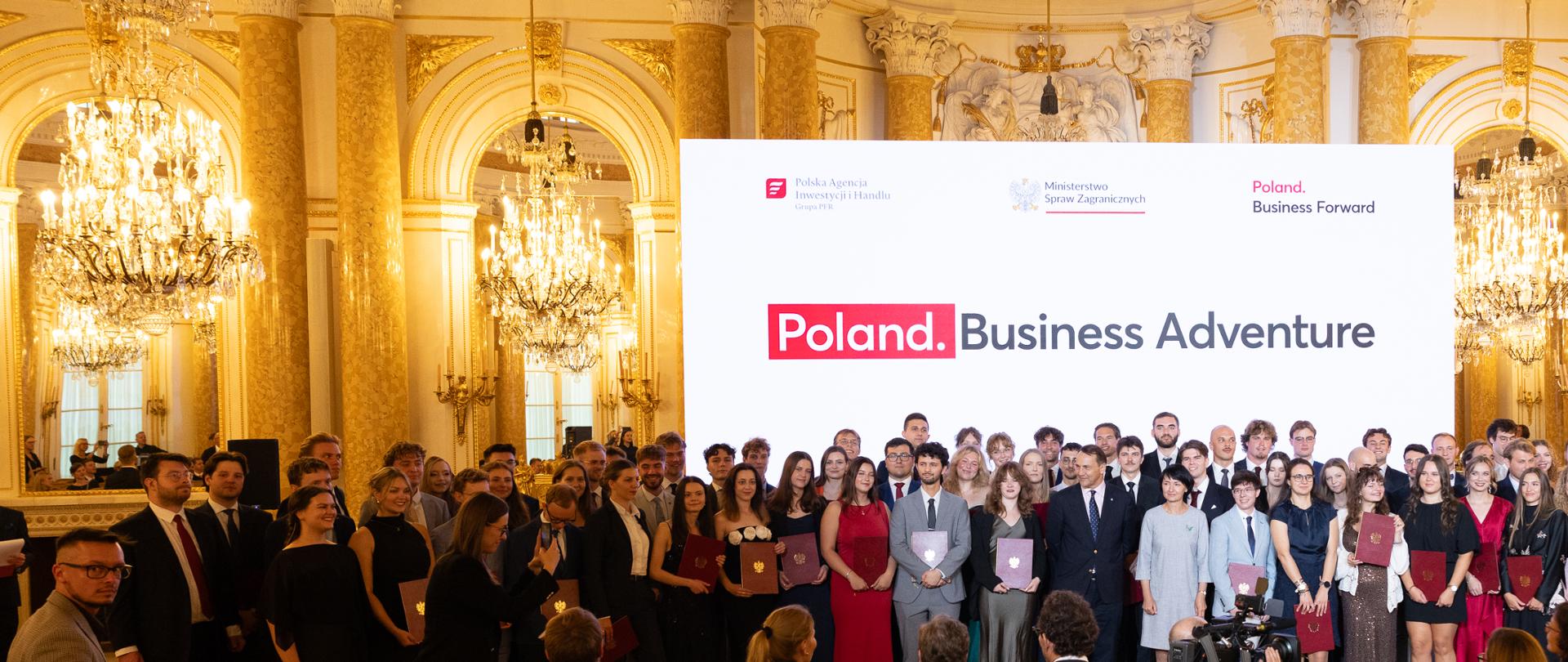 The photo shows a group of several dozen people. They are standing side by side and looking straight ahead. Behind them is a white wall with the words “Poland. Business Adventure.” Next to the wall are golden columns and chandeliers. 