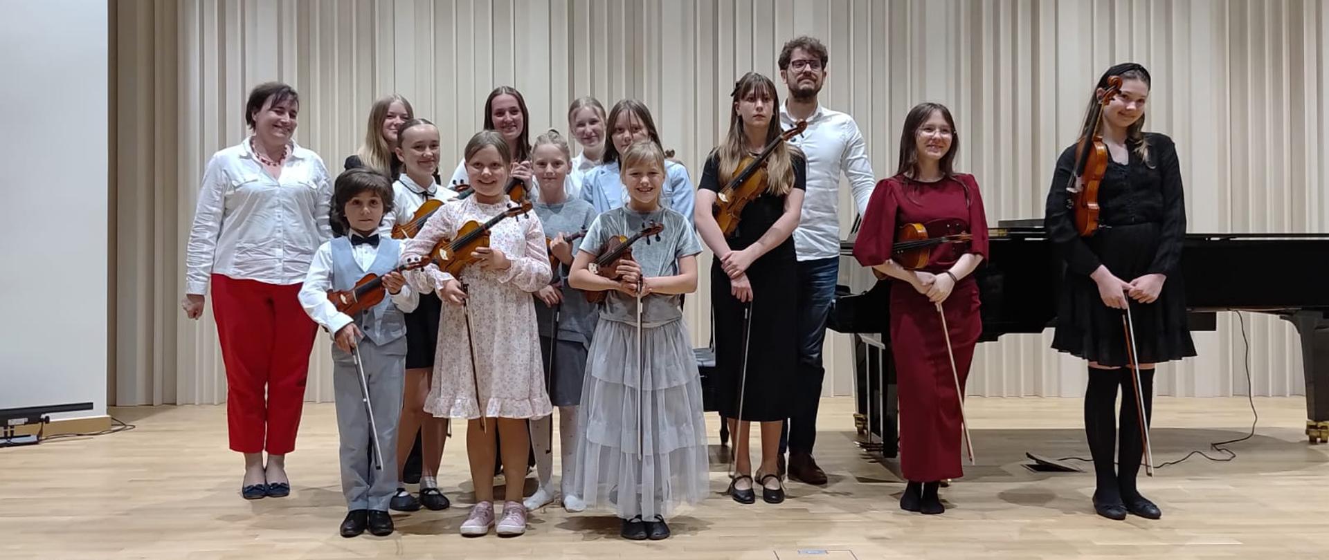 Na zdjęciu grupa odświętnie ubranych uczennic, które trzymają skrzypce. Po lewej stronie kobieta brunetka w czerwonych spodniach i białej koszuli. W tle za uczniami mężczyzna w białej koszuli, okularach i granatowych spodniach. Obok niego czarny fortepian. Tło obrazka w odcieniach beżu. 