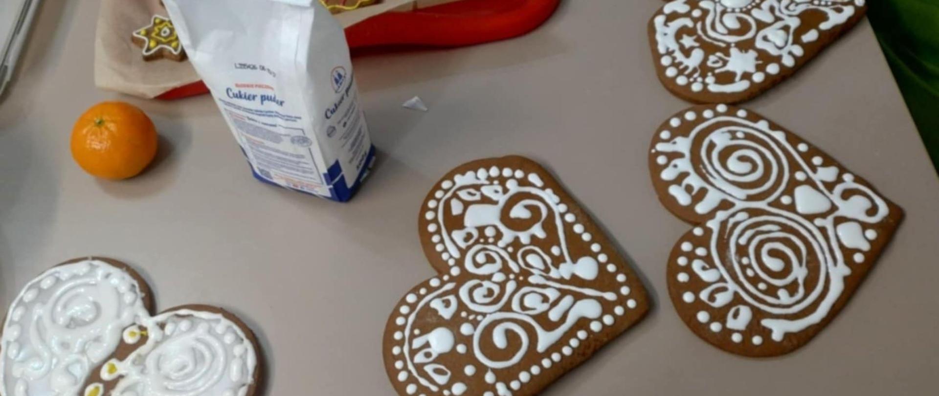 zdjęcie stołu na którym znajdują się cztery pierniki w kształcie serca ozdobione białym lukrem, obok torebka z cukrem, pomarańcza. 