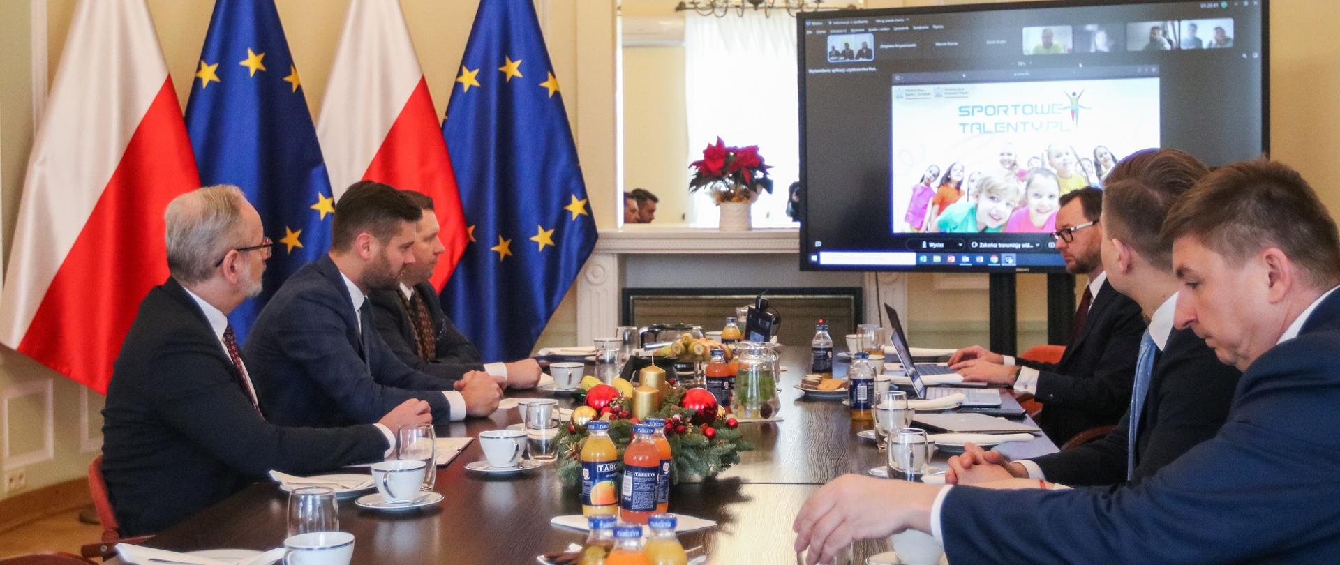 Przy długim stole siedzi kilka osób, za stołem wielki monitor, z drugiej strony flagi Polski i UE.