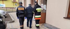 Zdjęcie przedstawia dwóch strażaków Ochotniczej Straży Pożarnej z Kamieńca pomagających mężczyźnie w dotarciu do punktu szczepień. W tle widać otwarte drzwi do punktu szczepień, samochody na parkingu oraz budynek.