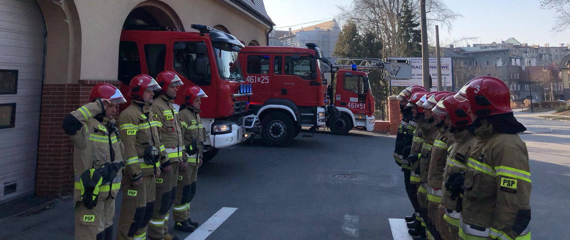 Plac przed budynkiem Komendy. Po lewej stronie widok na bramy garażowe. Przed bramami w szeregu stoją naprzeciw siebie strażacy w umundurowaniu specjalnym.