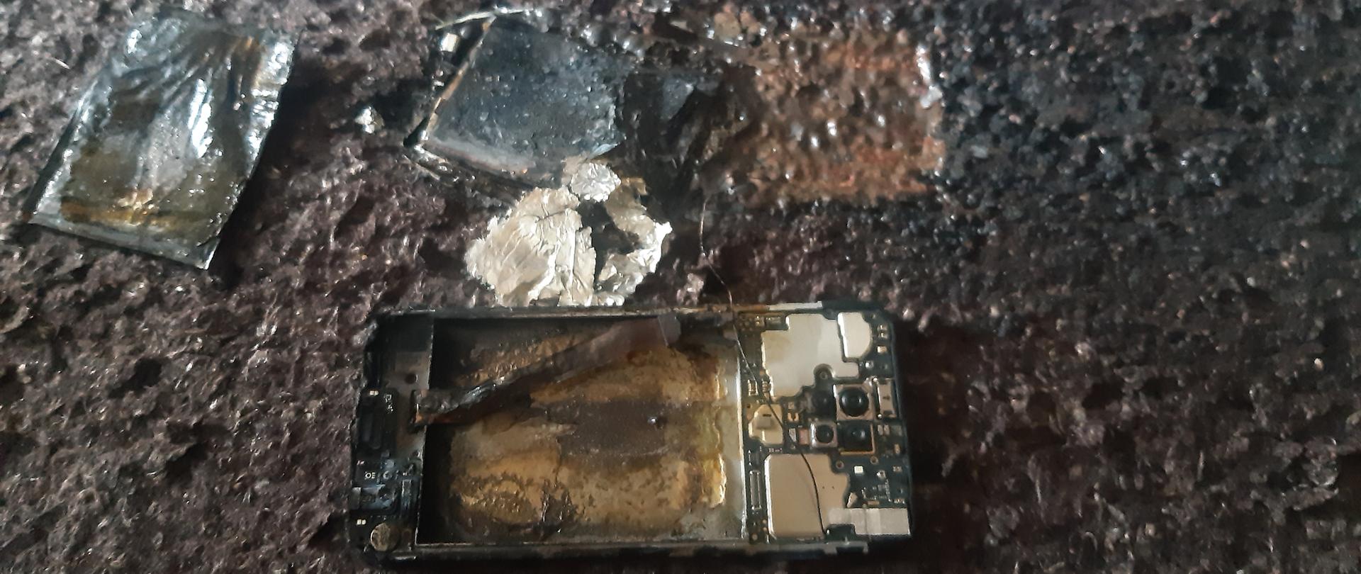 Zdjęcie przedstawia spalone elementy telefonu komórkowego leżące na nadpalonym dywanie