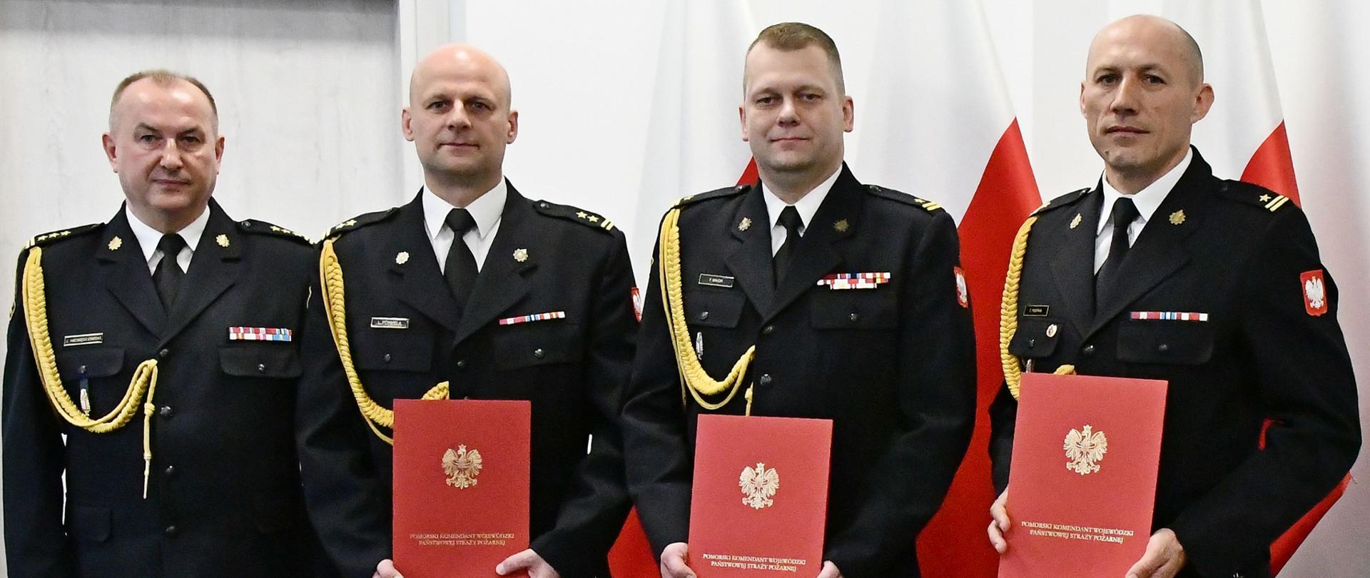 Czterech strażaków Państwowej Straży Pożarnej w mundurach wyjściowych ze sznurem stoi obok siebie trzech strażaków trzyma czerwone teczki za nimi ustawione są trzy flagi Polski oraz znajdują się drzwi. 