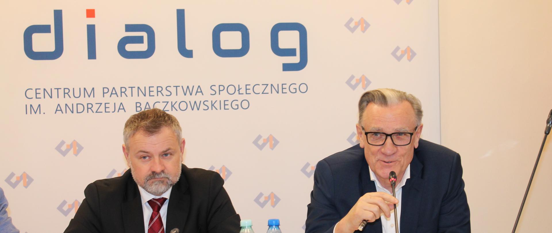 Na zdjęciu widnieje stół prezydialny za którym siedzą: Pan Grzegorz Wrona, Podsekretarz Stanu w Ministerstwie Aktywów Państwowych oraz Pan Jacek Kaczorowski, Współprzewodniczący Zespołu Trójstronnego ds. Branży Węgla Brunatnego ze strony pracodawców. W tle znajduje się baner z napisem Centrum Partnerstwa Społecznego „Dialog” im. Andrzeja Bączkowskiego