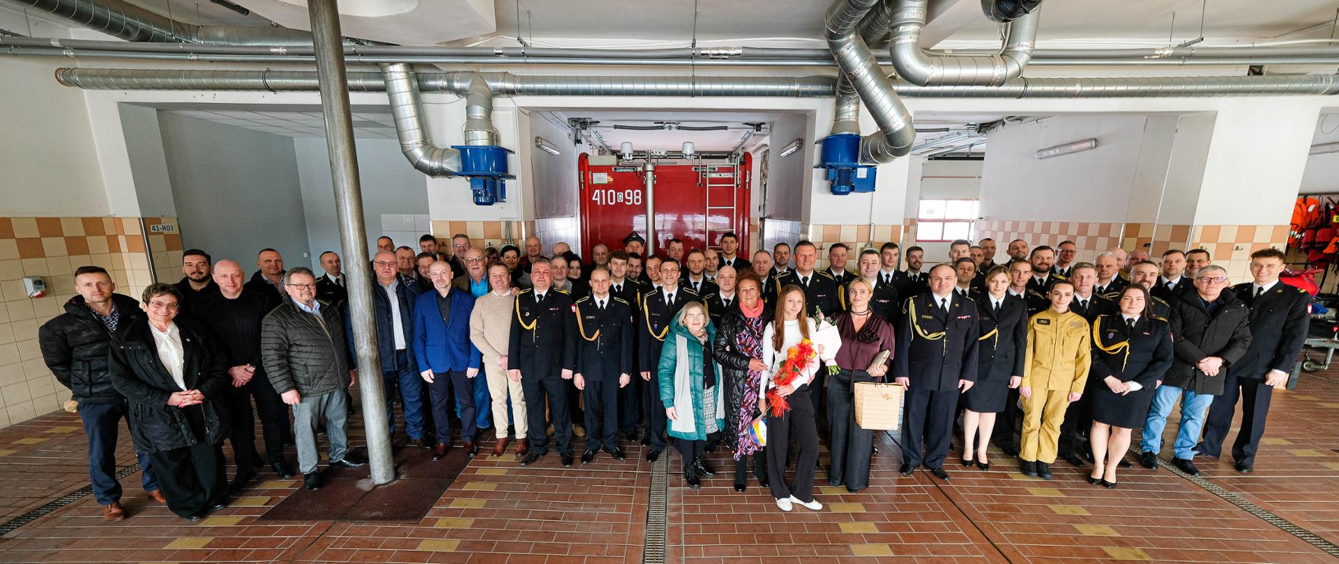 Zdjęcie grupowe uczestników uroczystości w garażu Jednostki Ratowniczo-Gaśniczej KP PSP w Chojnicach – strażacy, pracownicy oraz zaproszeni goście pozują do wspólnej fotografii.
